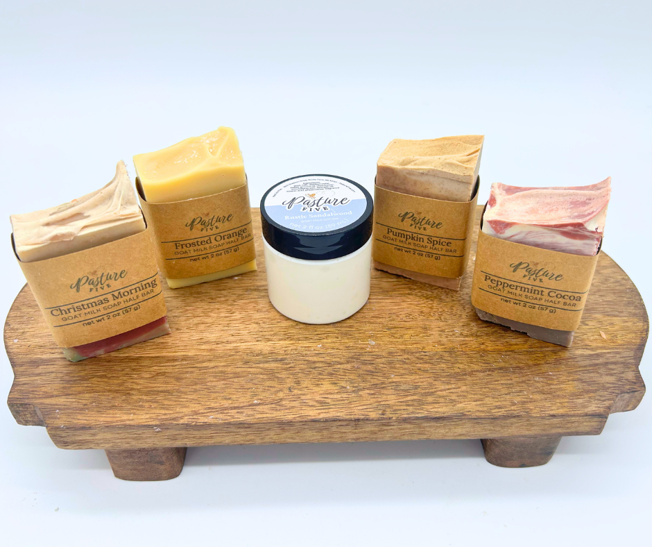 Four goat milk soaps and one jar of lotion, displayed on a rustic wooden stand against a clean white background.