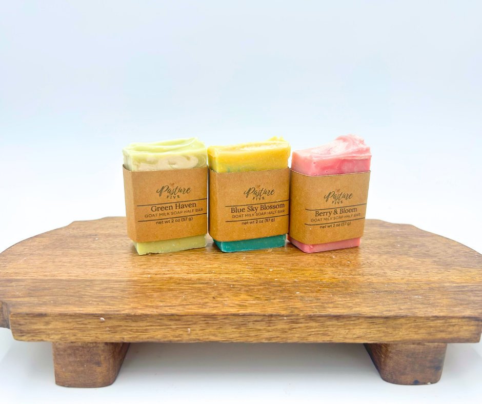 Three bars of soap on a wooden block with brown paper packaging against a light blue background.