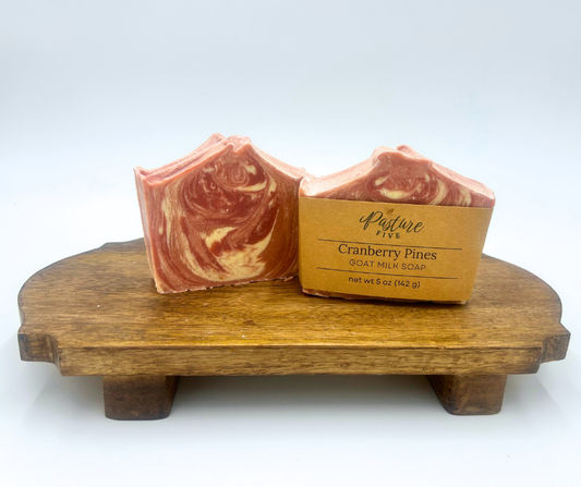 Two Cranberry Pine Goat Milk Soaps with a swirl design on a wooden stand with a white background.