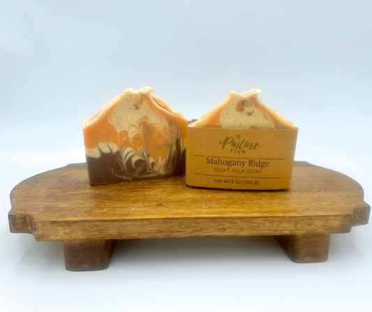 Two bars of goat milk soap with a swirl design on a wooden stand with a box labeled 'Pasture Five' on a white background.