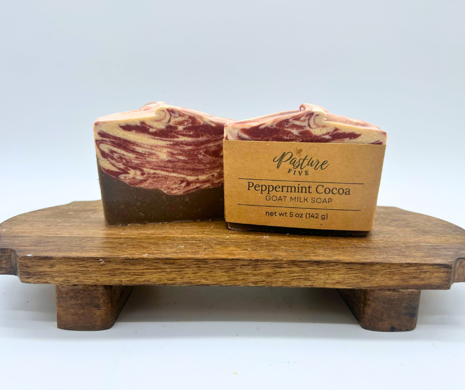 Two bars of marbled Peppermint Cocoa goat milk soap displayed on a small wooden stand against a light gray background.