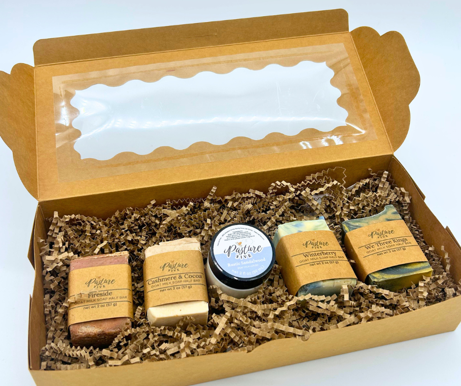 A gift box set containing four goat milk soaps and a small jar of goat milk lotion  in brown crinkle paper filler inside an open kraft cardboard box with a clear window lid.