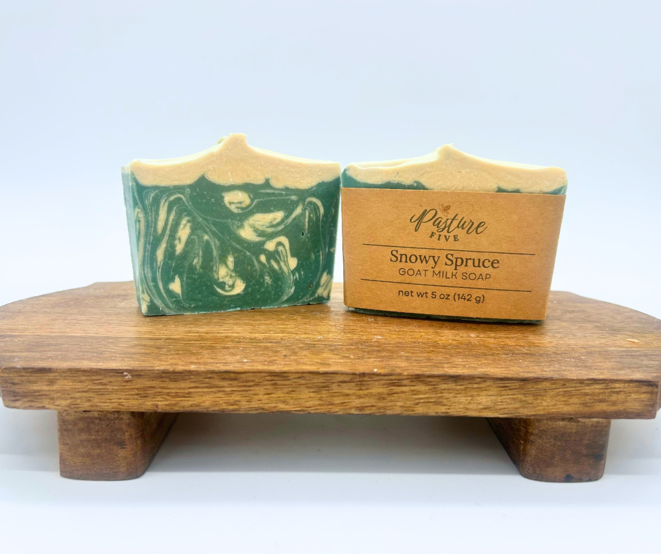 Two bars of Snowy Spruce goat milk soap bars, one with a wrap. Soaps are both on wood board. 