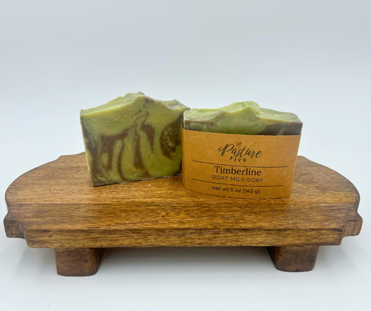 Two green and brown soap bars on a wooden stand with a label reading 'Pasture Five Timberline Goat Milk Soap'.