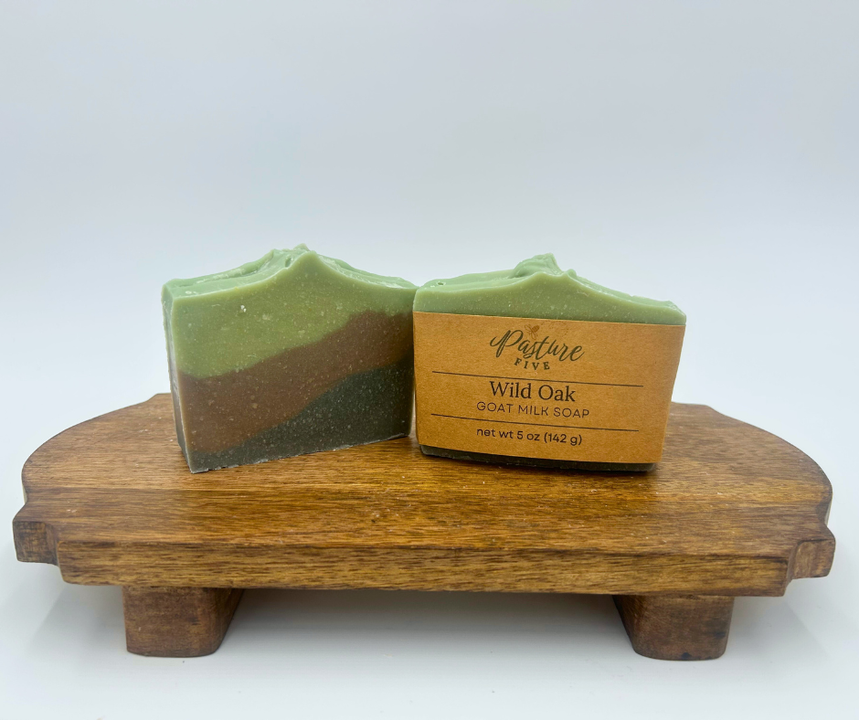 Two bars of soap on a wooden stand with a brown box labeled 'Pasture Five Wild Oak Soap'.