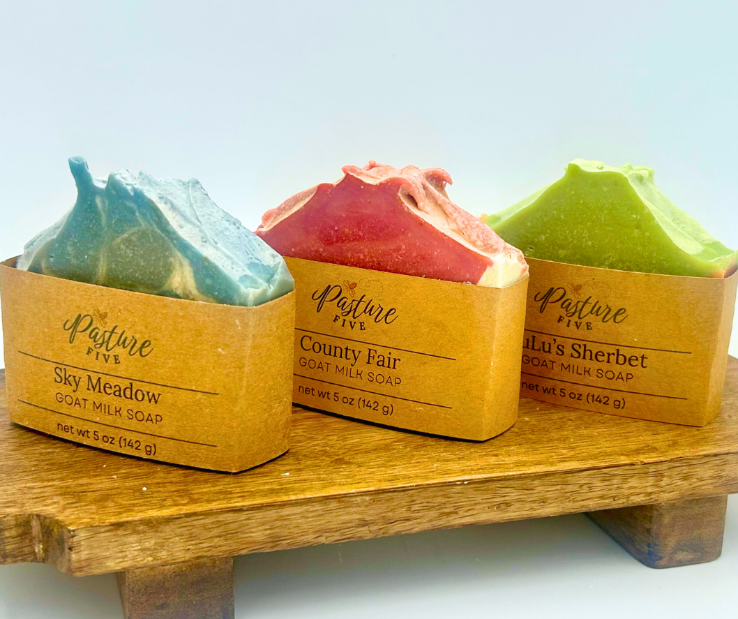 Three goat milk soaps in packaging on a wooden stand with a light blue background.