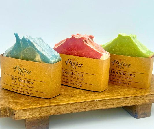 Three goat milk soaps in packaging on a wooden stand with a light blue background.